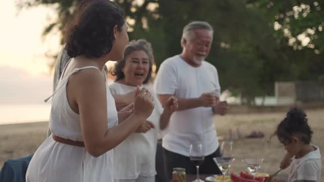 快乐的亚洲家庭在海滩度假。一群多代同堂的家庭在夏日日落的海滩上享受晚餐聚会和跳舞。小女孩与父母和祖父母一起放松和快乐。视频素材