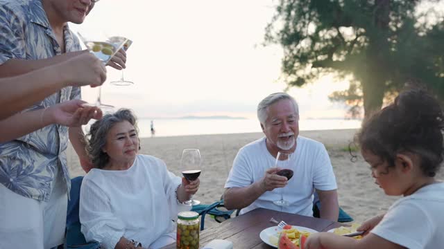 快乐的亚洲家庭在海滩度假。一群多代同堂的家庭在夏日日落的海滩上享受晚餐聚会和跳舞。小女孩与父母和祖父母一起放松和快乐。视频素材