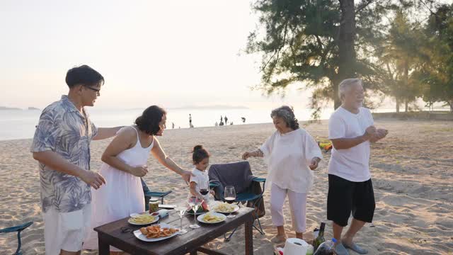 快乐的亚洲家庭在海滩度假。一群多代同堂的家庭在夏日日落的海滩上享受晚餐聚会和跳舞。小女孩与父母和祖父母一起放松和快乐。视频素材