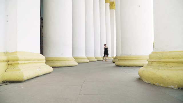 极宽的镜头优雅苗条的年轻芭蕾舞演员踮着脚尖在巨大的白色柱子之间跳舞。美丽的白人女人穿着黑色的衣服在户外表演。艺术和表演概念。视频素材