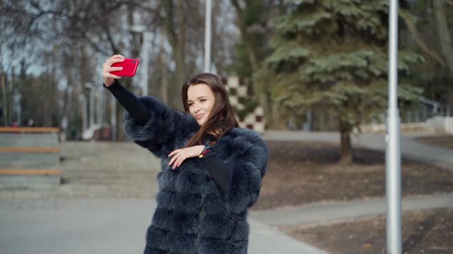 一个漂亮女孩在公园自拍的照片。年轻的女人在一个阳光明媚的冬天的红色手机上做豪华皮草大衣的照片。视频素材