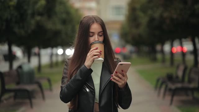 一位年轻女子正在散步。她一边用智能手机发短信，一边喝咖啡。4 k。视频素材