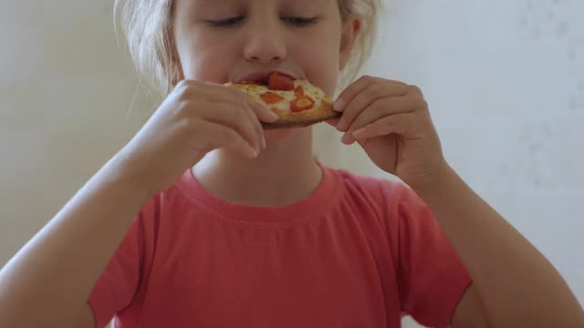 孩子吃披萨。饥饿的小女孩坐在厨房的餐桌上吃着美味的意大利披萨。不健康的营养快餐。一个吃午饭的孩子的肖像。面对年轻女孩特写。视频素材