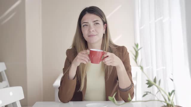 肖像梦幻体贴美丽的女人闻美味的咖啡或茶在杯子和微笑。年轻自信的白人黑发女士坐在咖啡馆思考。生活方式和美容理念。视频素材