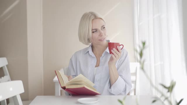 幸福美丽的金发女人的肖像坐在自助餐厅与书和咖啡杯看着镜头微笑。美丽的白人女士享受室内休息。爱好和审美观念。视频素材