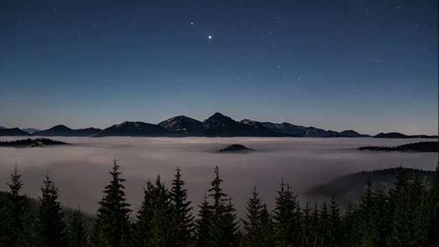 蓝色的夜空与星星在高山之上，森林和雾云在月光下快速移动傍晚自然景观时间流逝视频素材