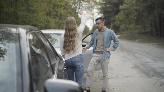 年轻的男人和女人在路边一边大喊一边打手势。愤怒的白人千禧一代司机在车祸室外争吵。道路冲突和汽车破损。视频素材