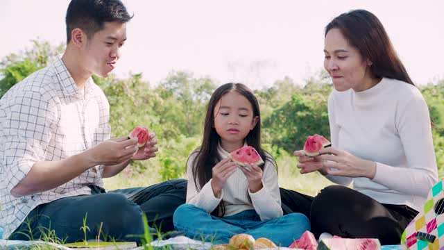 幸福的家庭野餐。亚洲父母(父亲，母亲)和小女孩吃西瓜，有乐趣和享受自己一起在野餐布在花园在一个阳光明媚的日子视频素材