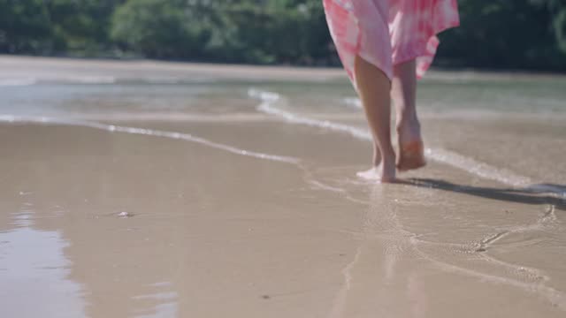 女赤脚走在岛上的海滩上，清澈的海水海浪在缓慢的动作，溅水打腿。后视图低摄像角度，夏季旅游预订，热带气候国家视频素材