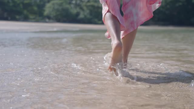 女性的腿和脚沿着岛屿的海滩与溅水的波浪溅水和赤脚在慢动作视频素材