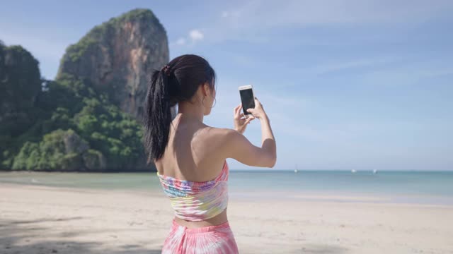 年轻的亚洲女士拍美丽的海洋风景，暑假旅行，女性游客享受岛屿海滩，使用智能手机拍照的假期记忆，阳光海景海滩前视频素材