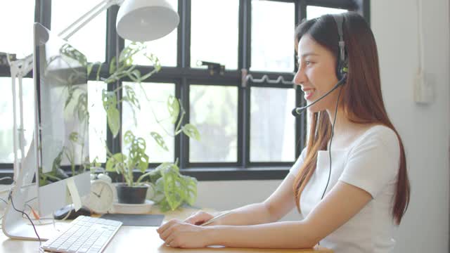 亚洲女学生戴着无线耳机与skype老师在线学习，快乐的年轻女子学习语言听讲座看网络研讨会写笔记看笔记本电脑坐在家里学习视频素材