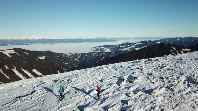 无人机拍摄到一对夫妇穿着雪地鞋徒步前往Amerinkogel峰。奥地利阿尔卑斯山的冬天。无尽的山脉，覆盖着粉状的雪。山谷里有浓雾。户外活动视频素材