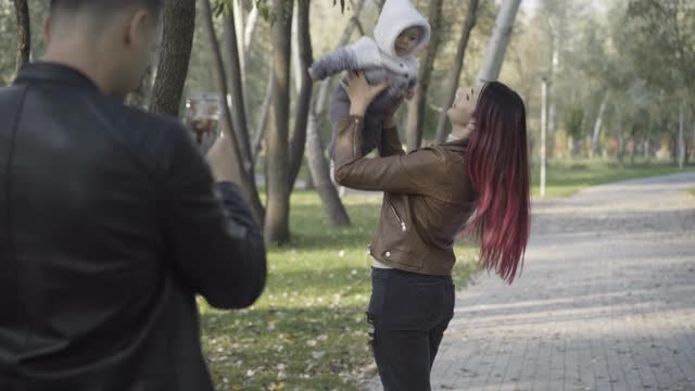 年轻的白人男子拍照微笑美丽的女人与婴儿在阳光灿烂的公园。丈夫和父亲与妻子和儿子一起享受周末户外活动。家庭的概念视频素材