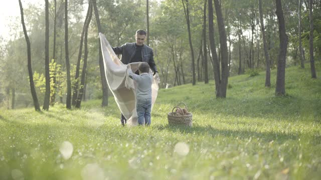 广角镜头，白种人年轻的父亲和小儿子躺在绿色的夏季草地上的毯子。快乐的男人和男孩在阳光明媚的户外家庭野餐。休闲幸福观视频素材