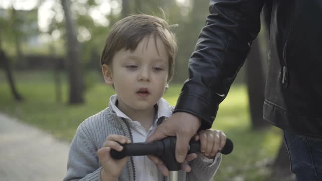 快乐的白人小男孩骑摩托车在阳光与年轻男性的手拉方向盘。陌生的父亲和可爱的儿子在春天或秋天的公园散步视频素材