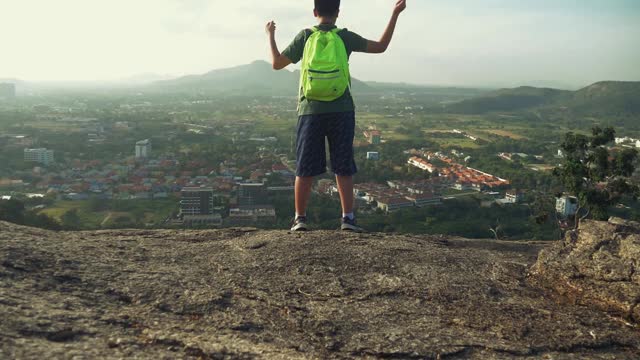 背包男孩站在山顶举起双手，在早晨看风景，积极的生活理念。视频素材