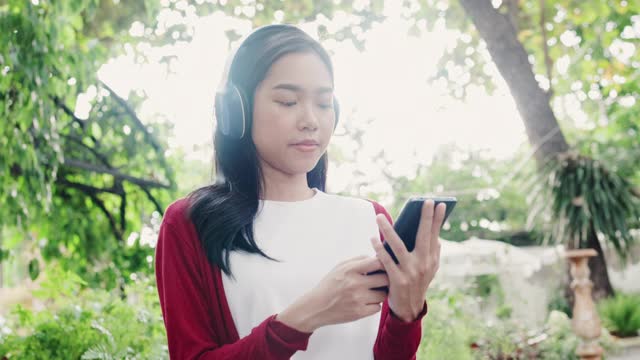 美丽的亚洲女人戴着耳机在房子的花园里听着快乐的音乐。放松，假期自由自在。好天气遮天蔽日。呆在家里的概念视频素材