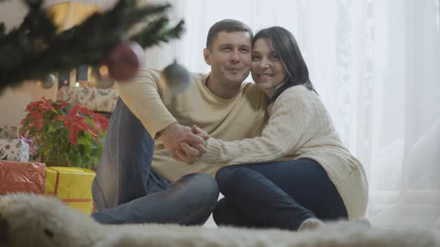 幸福的白人夫妇坐在家里的软地毯上，圣诞树和大笑。积极的成年男人和女人在室内享受新年除夕的肖像。假期季节的概念视频素材