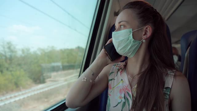 女乘客戴着保护面罩，在当地的火车上开车，用手机聊天，女人看着窗外，微笑着路过火车站。全球冠状病毒大流行期间的运动和生活方式视频素材