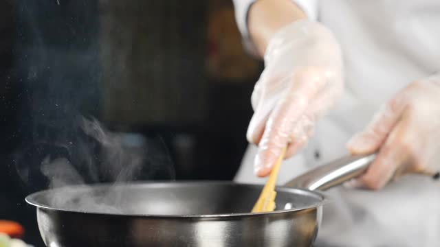 在餐厅做饭。慢动作视频。女厨师戴着手套，穿着白色制服，用平底锅里的桨搅拌食物。传统的地中海式饮食。全高清视频素材