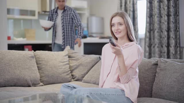 积极的年轻女子切换遥控器作为男人来与爆米花。轻松愉快的白人夫妇在家里的客厅里看电视。休闲和生活方式的概念。视频素材