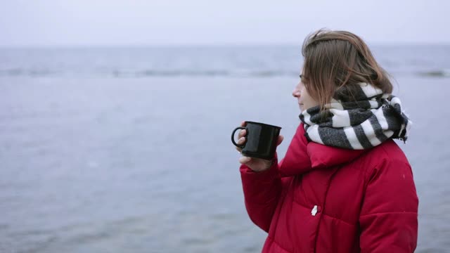 一位年轻女子端着一杯茶，欣赏着冬日的风景。视频素材