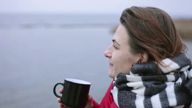 年轻女子捕捉雪花。一个女人端着一杯热茶站在湖边。视频素材