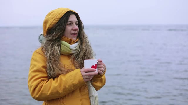 冬天，年轻女子在一个大湖的岸边喝着热茶。视频素材