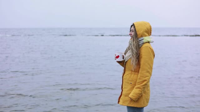 一个女人站在一个大湖的岸边，手里拿着一杯热茶。视频素材