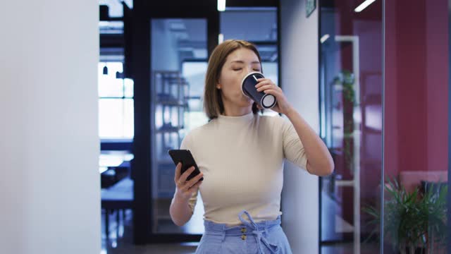 在现代办公室里拿着咖啡杯拿着智能手机走路的亚洲女商人视频素材