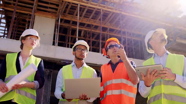 多民族的专家和中年工头使用数码平板电脑和笔记本电脑走下建筑工地，成熟的人使用定额与其他专家进行交谈视频素材