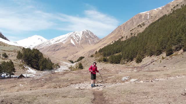 一个男人的鸟瞰图在帽子和短裤和一个背包与徒步杆子沿着高山山谷视频素材