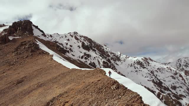 登山者在石头和雪的交界处的山脊上，背着背包和拐杖爬上山坡。北高加索山脉高处鸟瞰图视频素材