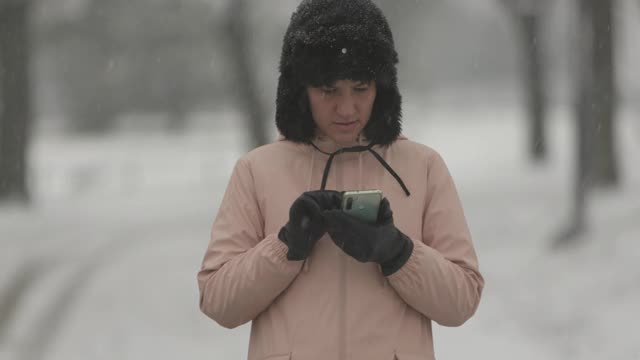 戴着帽子和手套的女人在下雪和打电话时穿过公园视频素材