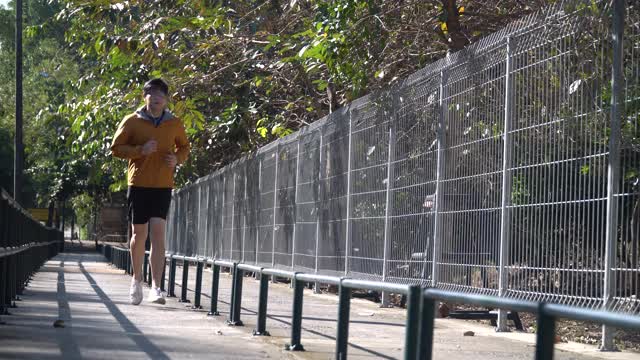 亚洲年轻的跑步者男子穿着毛衣兜帽听音乐在公园在早上在户外。适合男性慢跑锻炼。运动训练视频素材