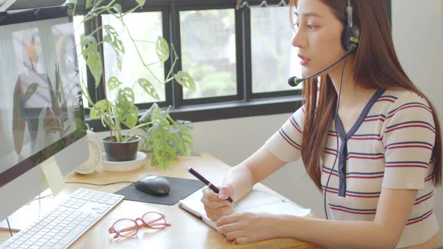 亚洲女学生戴着无线耳机与skype老师在线学习，快乐的年轻女子学习语言听讲座看网络研讨会写笔记看笔记本电脑坐在家里学习视频素材