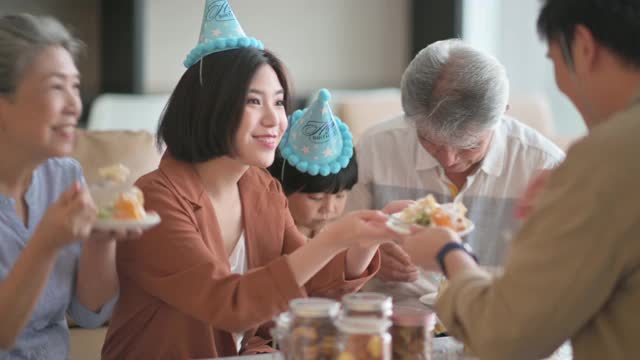 亚洲华人多代同堂庆祝生日，孙子戴着派对帽在客厅吃生日蛋糕视频素材