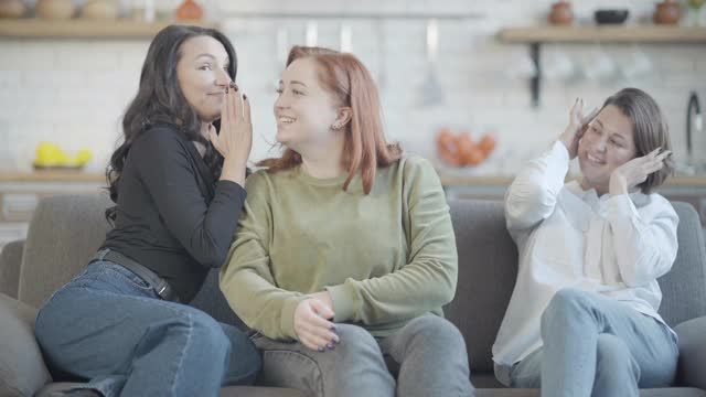 快乐苗条的女人对着大号红发朋友的耳朵窃窃私语，微笑的女人坐在沙发上闭着耳朵。积极友好的白人女性在室内分享秘密。视频素材