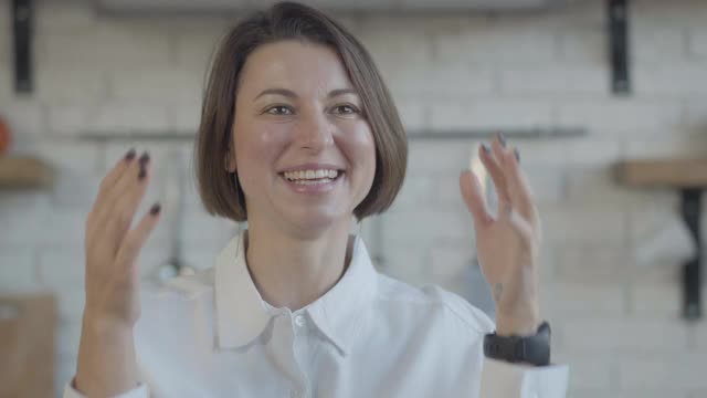 愉快的褐发年轻白种人女人大笑和触摸脸的特写。积极快乐的女士坐在厨房在家里的乐趣。休闲的生活方式。视频素材