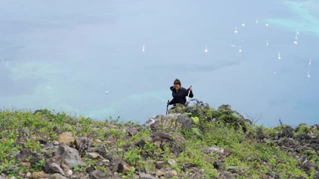 一个年轻女子徒步攀登陡峭的绿色山丘前的一个遥远的海湾与白色的小帆船-班达，印度尼西亚视频素材