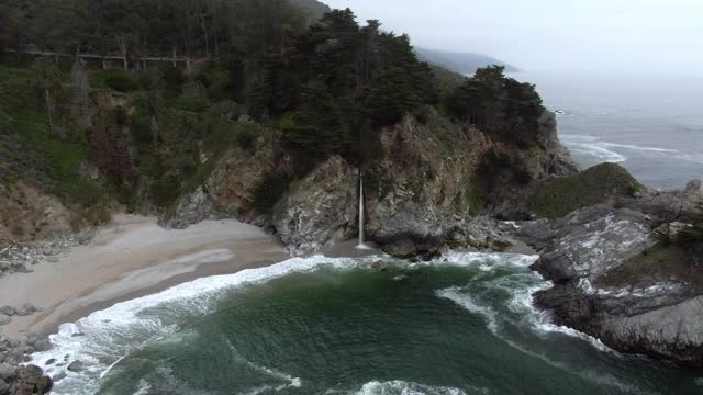 空中拍摄的瀑布从岩石溅在海滩上，无人机下降到瀑布对树-大苏尔，加利福尼亚视频素材