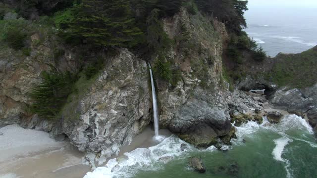 空中平移拍摄的瀑布从岩石在海滩溅水，无人机飞行瀑布对着树-大苏尔，加利福尼亚视频素材