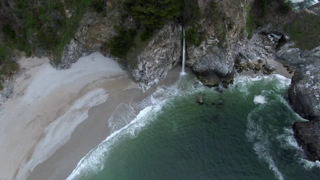 空中倾斜拍摄的瀑布从岩石溅在海滩上，无人机从瀑布向后下降对树-大苏尔，加利福尼亚视频素材