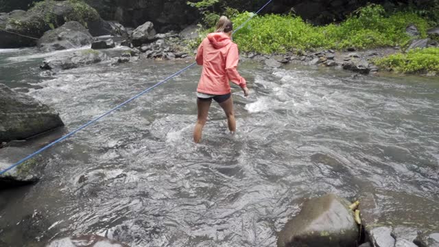 慢镜头锁定，一名女子徒步旅行者在向导绳的帮助下小心地穿过一条湍急的河流，四周都是岩石和热带植物——印度尼西亚的巴厘岛视频素材