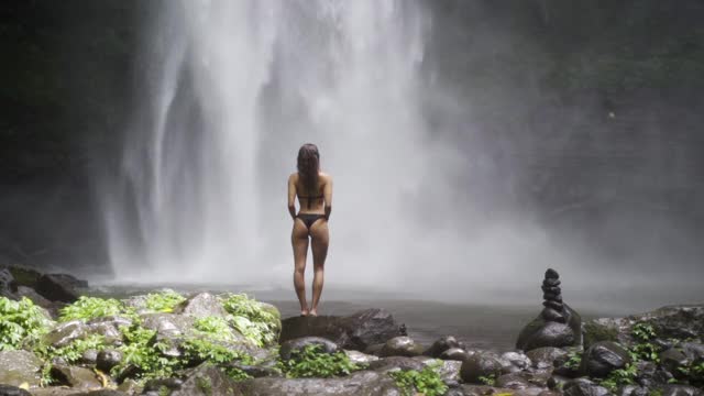 一名身穿比基尼的女子站在阳光照耀下的岩石上，面对湍急的野生瀑布——印尼巴厘岛视频素材