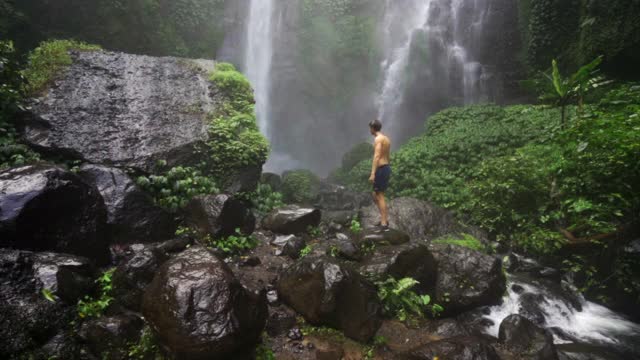 一个赤膊男子攀登的岩石路径与高奔流瀑布周围郁郁葱葱的热带植物覆盖悬崖-巴厘岛，印度尼西亚视频素材