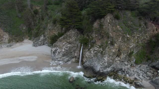 空中平移拍摄瀑布从岩石溅在海滩上，无人机飞瀑布对着树-大苏尔，加利福尼亚视频素材