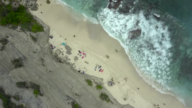 空中向后移动的海滩悬崖边与碰撞的海浪，绿松石的水，和日光浴者在阳光浸泡的沙子-努沙佩尼达和龙目岛，巴厘岛视频素材