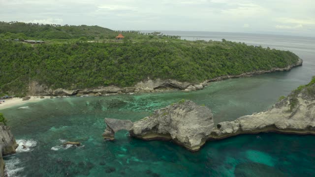 空中向前移动到一个拱形岩石形成的热带海滩海湾，绿松石的水，陡峭的海滩悬崖，珊瑚礁，和溅起的海浪-努沙佩尼达和龙目岛，巴厘岛视频素材